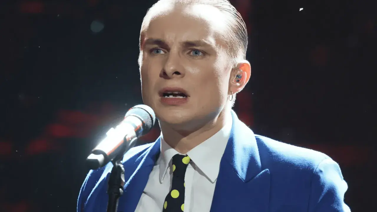 Ralph Kaminski, w niebieskiej marynarce i krawacie w grochy, śpiewa do mikrofonu. Jego partner, wokalista, jest w centrum uwagi.