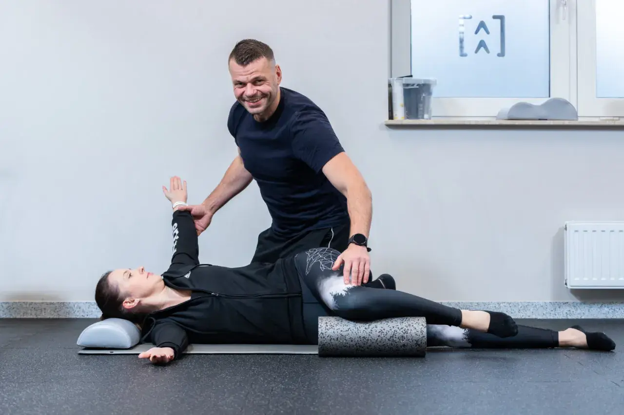 Fizjoterapeuta pomaga pacjentce w ćwiczeniach na kręgosłup. Kobieta leży na macie, z nogami ułożonymi na wałku, a mężczyzna koryguje jej pozycję ramienia.
