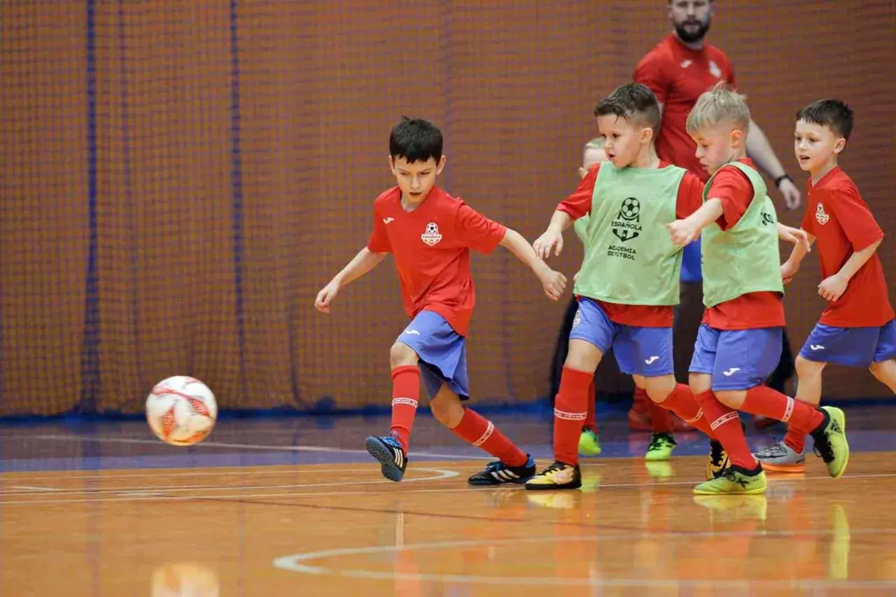 Zacięte współzawodnictwo sportowe dzieci i młodzieży na hali. Mali piłkarze w czerwonych koszulkach i zielonych kamizelkach walczą o piłkę.