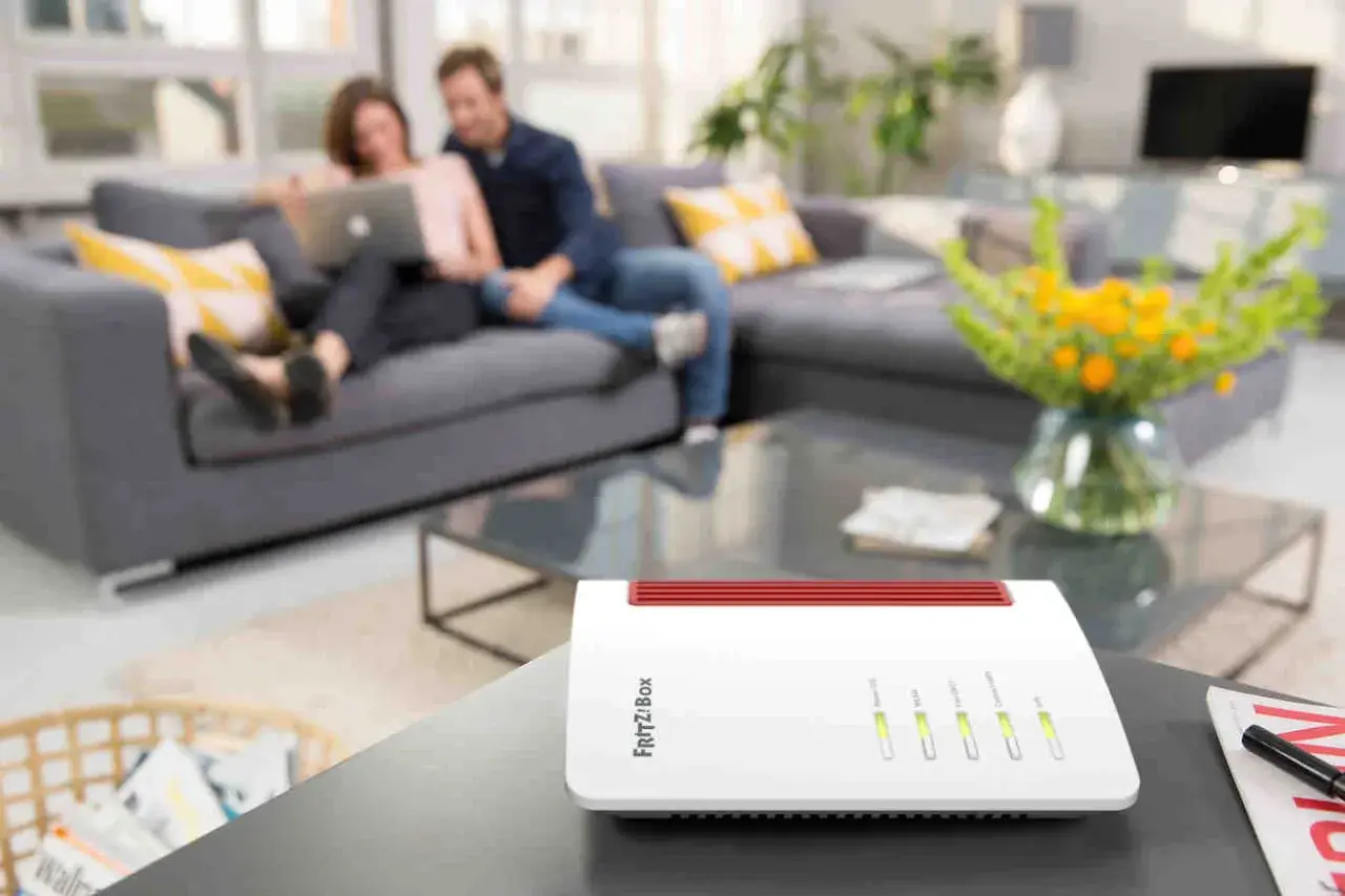 Biały router FRITZ!Box na stoliku, z parą relaksującą się na kanapie w tle. To urządzenie zapewnia domowe Wi-Fi.