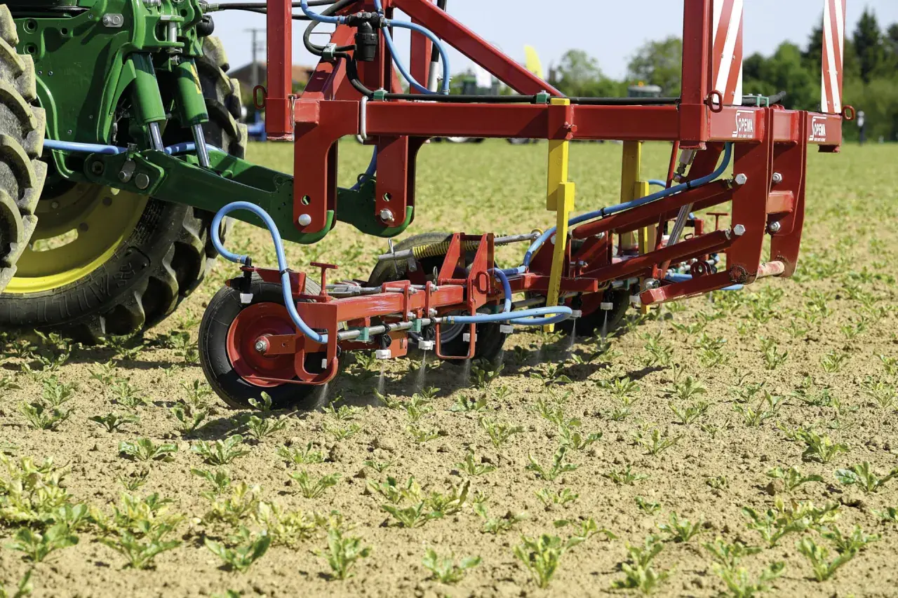Un tracteur vert et un pulvérisateur rouge travaillent dans un champ de jeunes pousses. La machine applique un traitement ciblé, peut-être pour une liste de graminées à éradiquer.