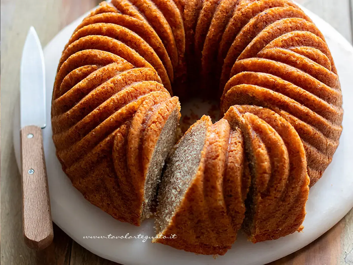 Una torta cachi e mele dorata, con un bordo scanalato, è affettata su un tagliere bianco. Un coltello con manico in legno è appoggiato accanto.
