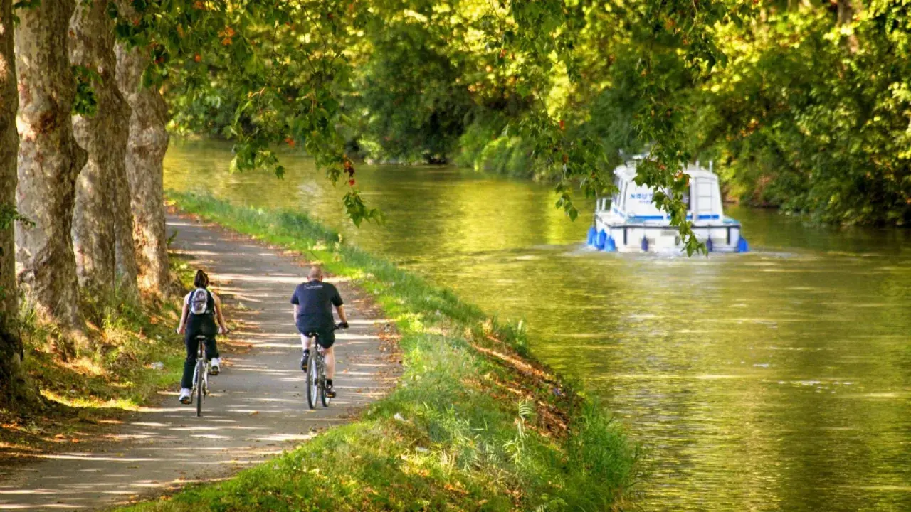 Pareja en bicicleta disfruta de un **canal du midi recorrido** bajo árboles frondosos, mientras una barcaza navega por el agua.