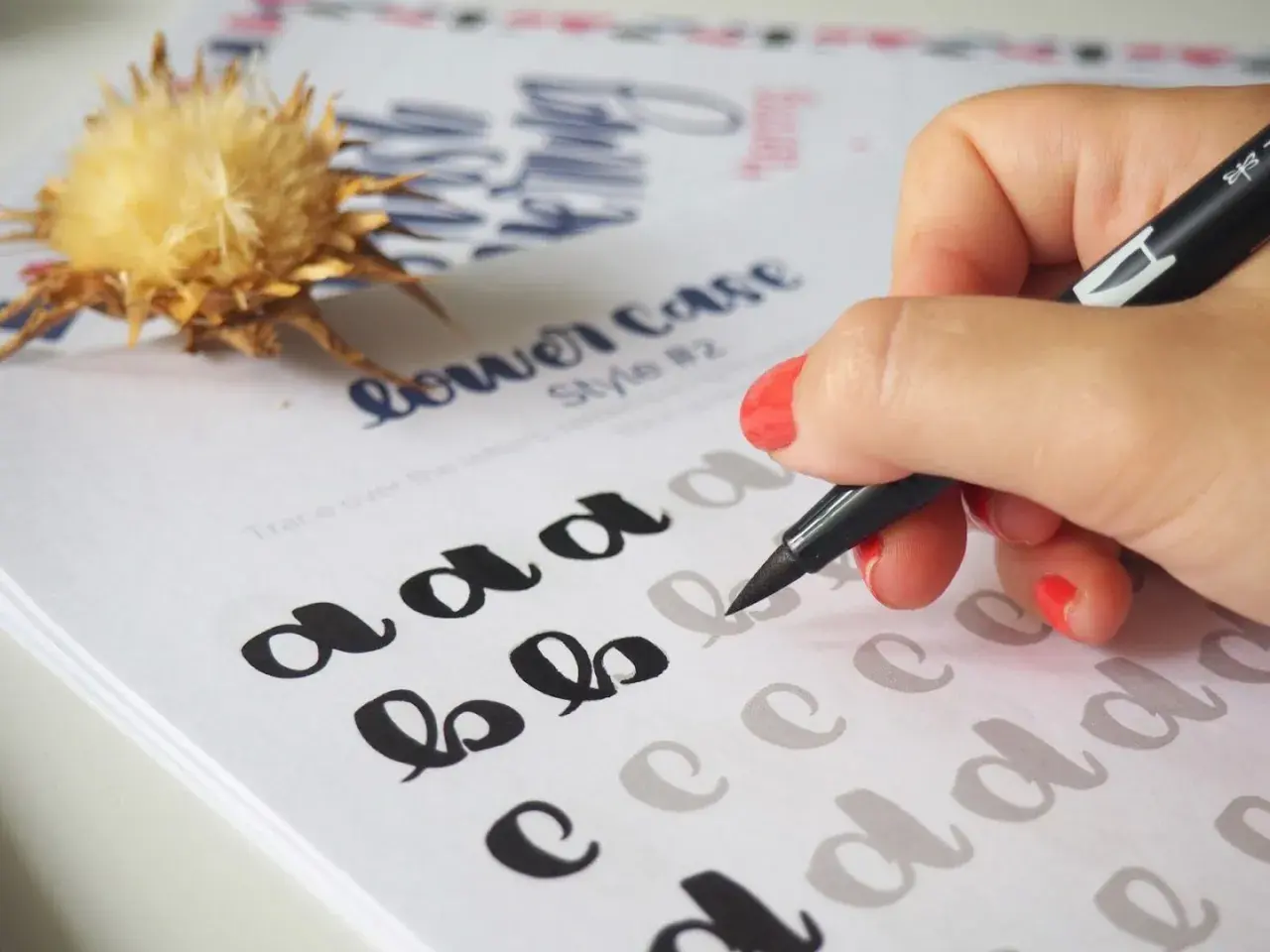 Mano practicando lettering con un rotulador negro sobre una hoja con letras "a", "b", "c" y "d" para calcar.