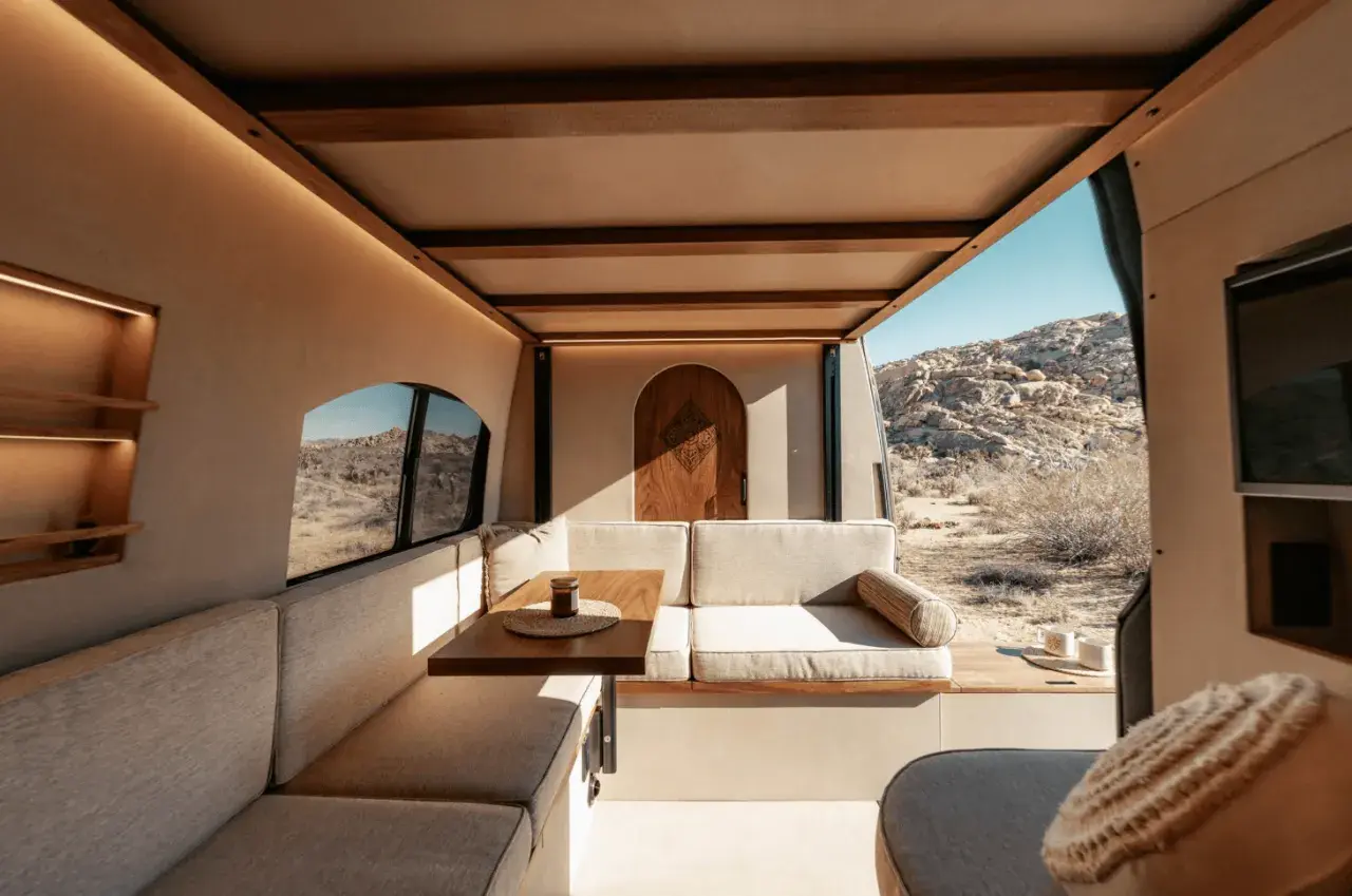 Intérieur de camping-car aménagé avec banquettes, table et porte en bois, vue sur un paysage désertique.
