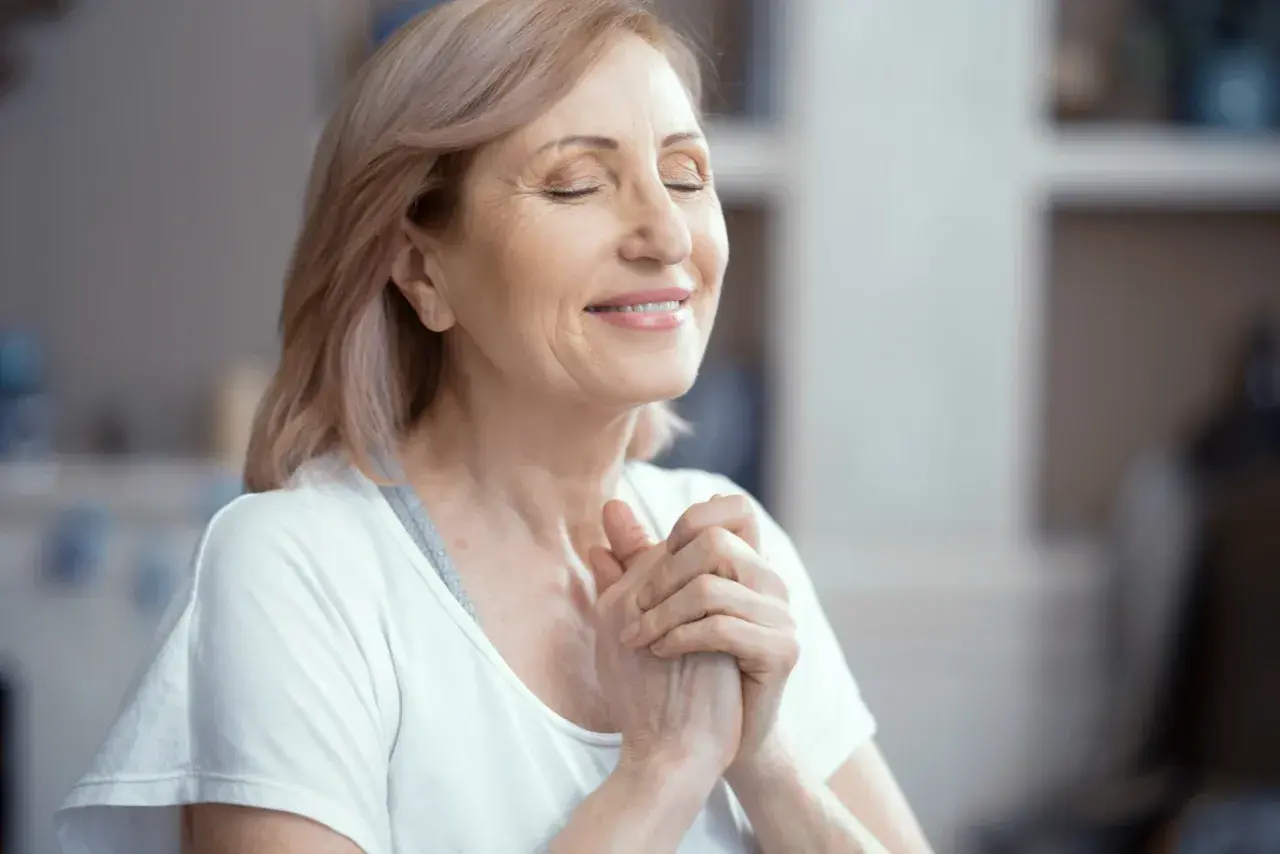 Uśmiechnięta kobieta z zamkniętymi oczami, dłonie złożone na piersi, praktykuje techniki mindfulness.