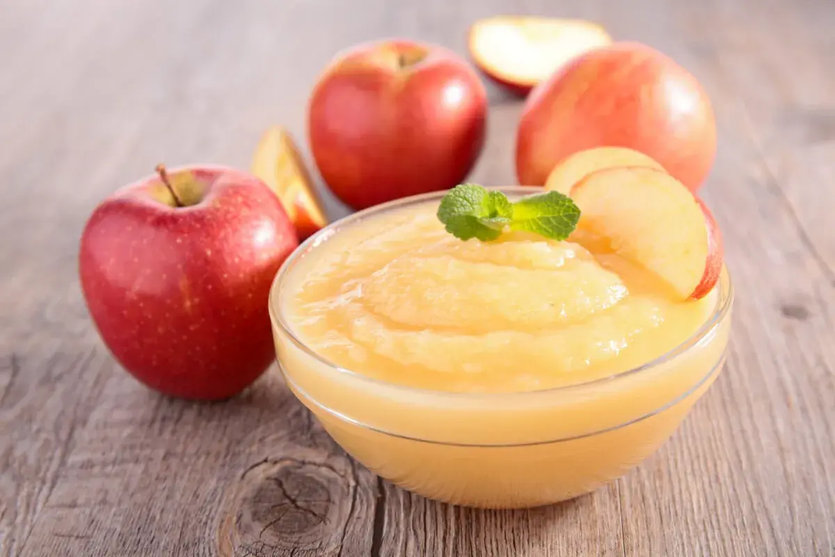 Un tazón de deliciosa salsa de manzana casera, decorado con menta y rodajas de manzana, rodeado de manzanas rojas frescas sobre una mesa de madera.
