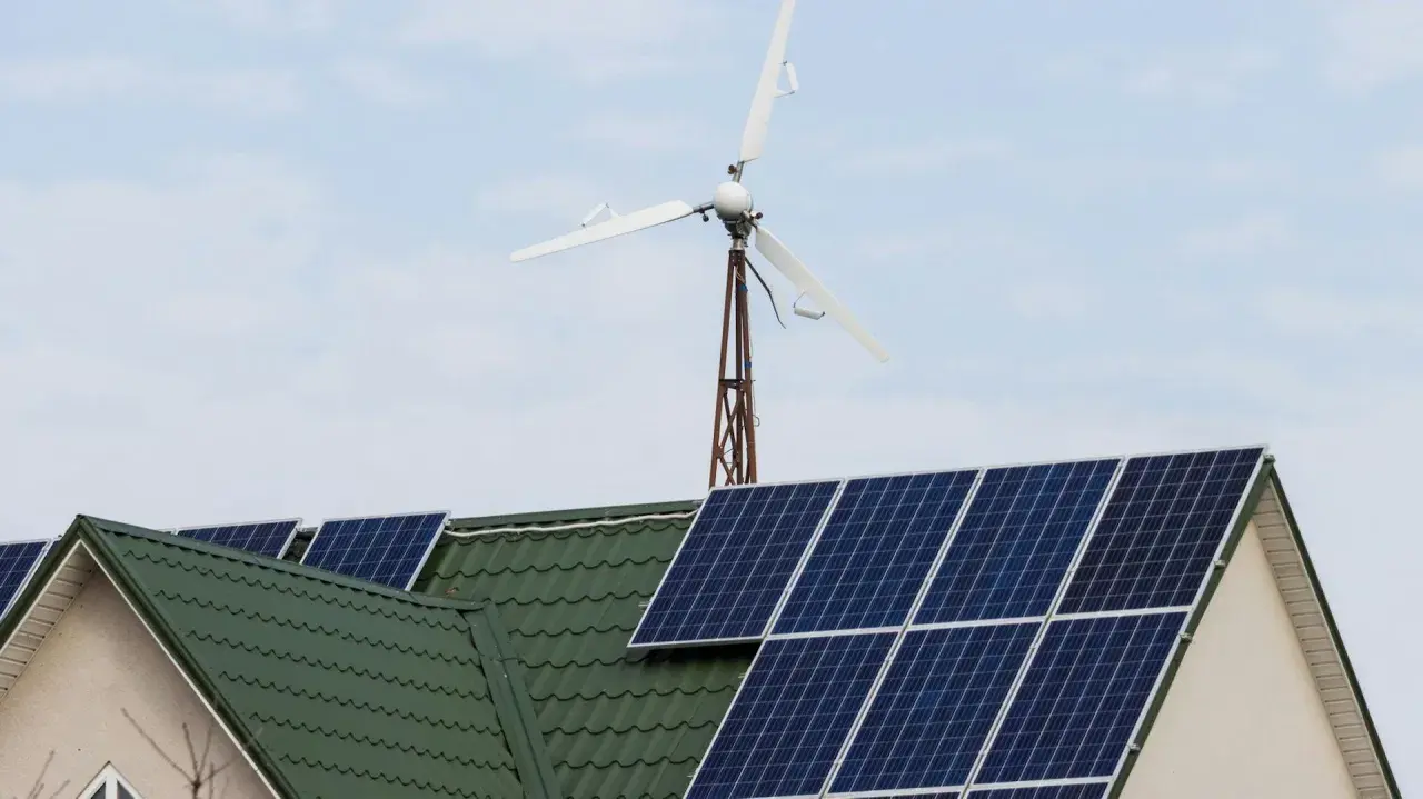 Panele słoneczne na zielonym dachu domu, nad nimi mała turbina wiatrowa.