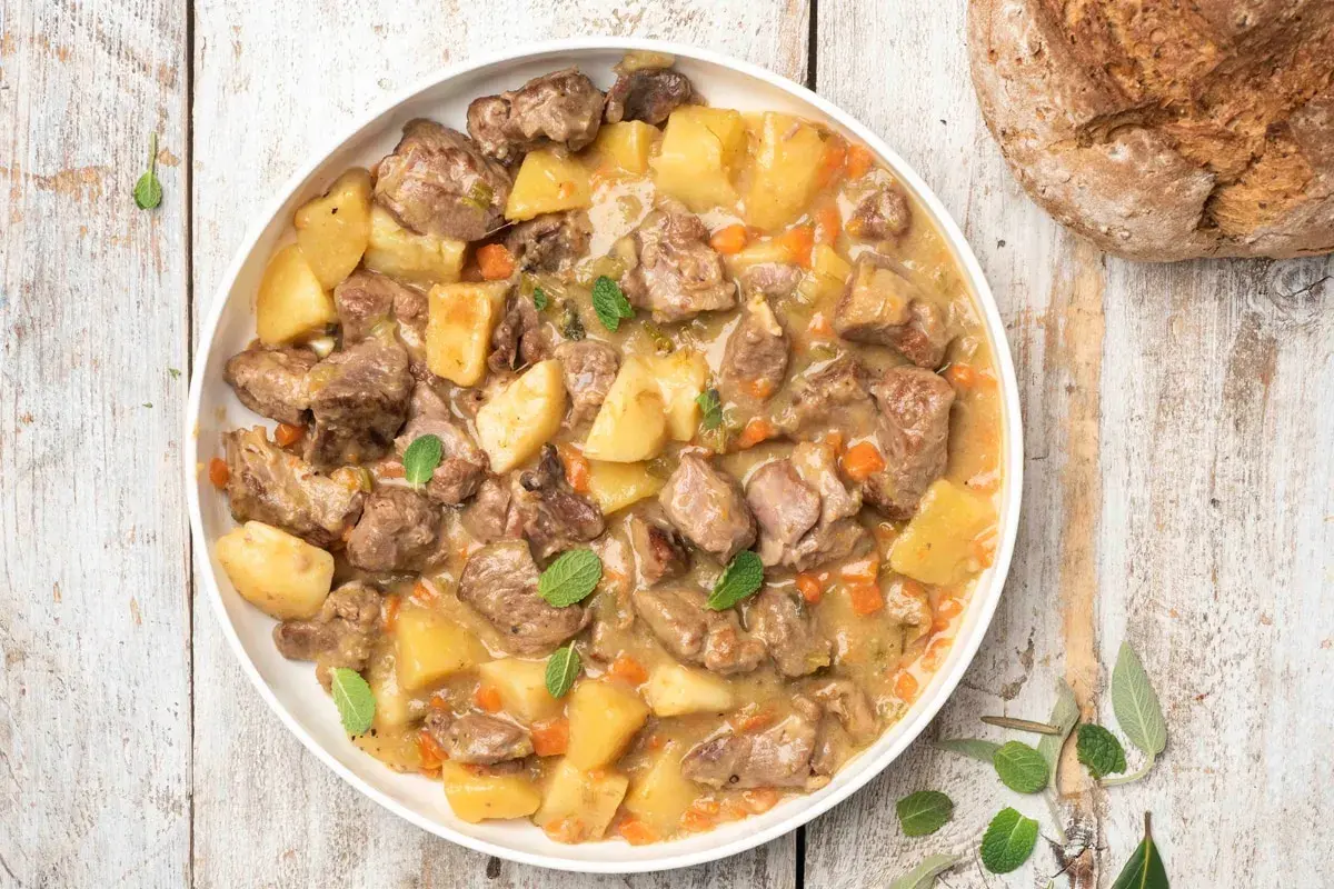 Un saporito agnello in umido con patate, carote e erbe aromatiche, servito in un piatto bianco su un tavolo di legno rustico.