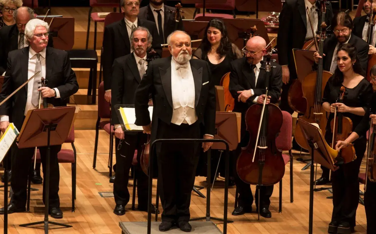 Orkiestra przygotowuje się do wykonania Dies Irae. Dyrygent w białej kamizelce i fraku stoi na podwyższeniu, otoczony muzykami z instrumentami smyczkowymi.