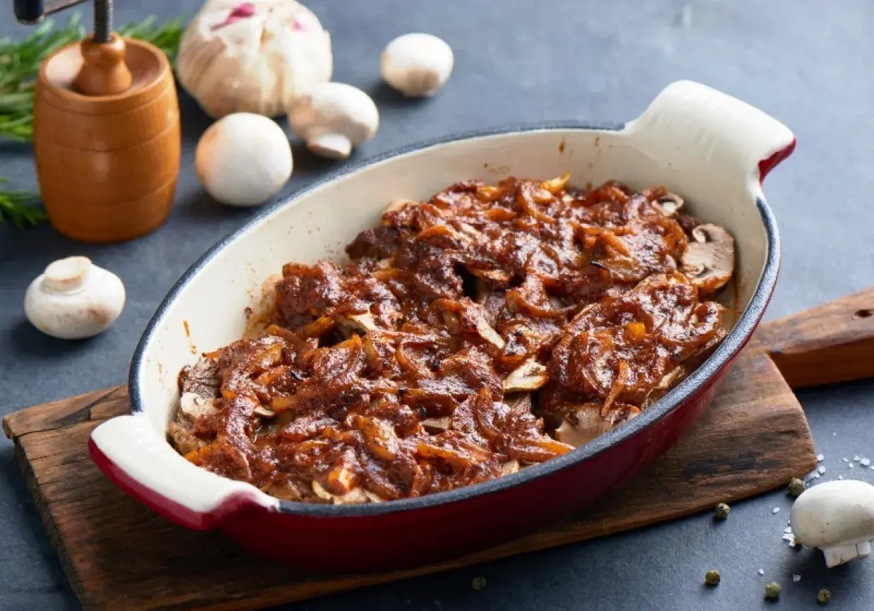 Karkówka pieczona w plastrach z cebulą i ketchupem, podana w naczyniu żaroodpornym. Obok pieczarki i czosnek.