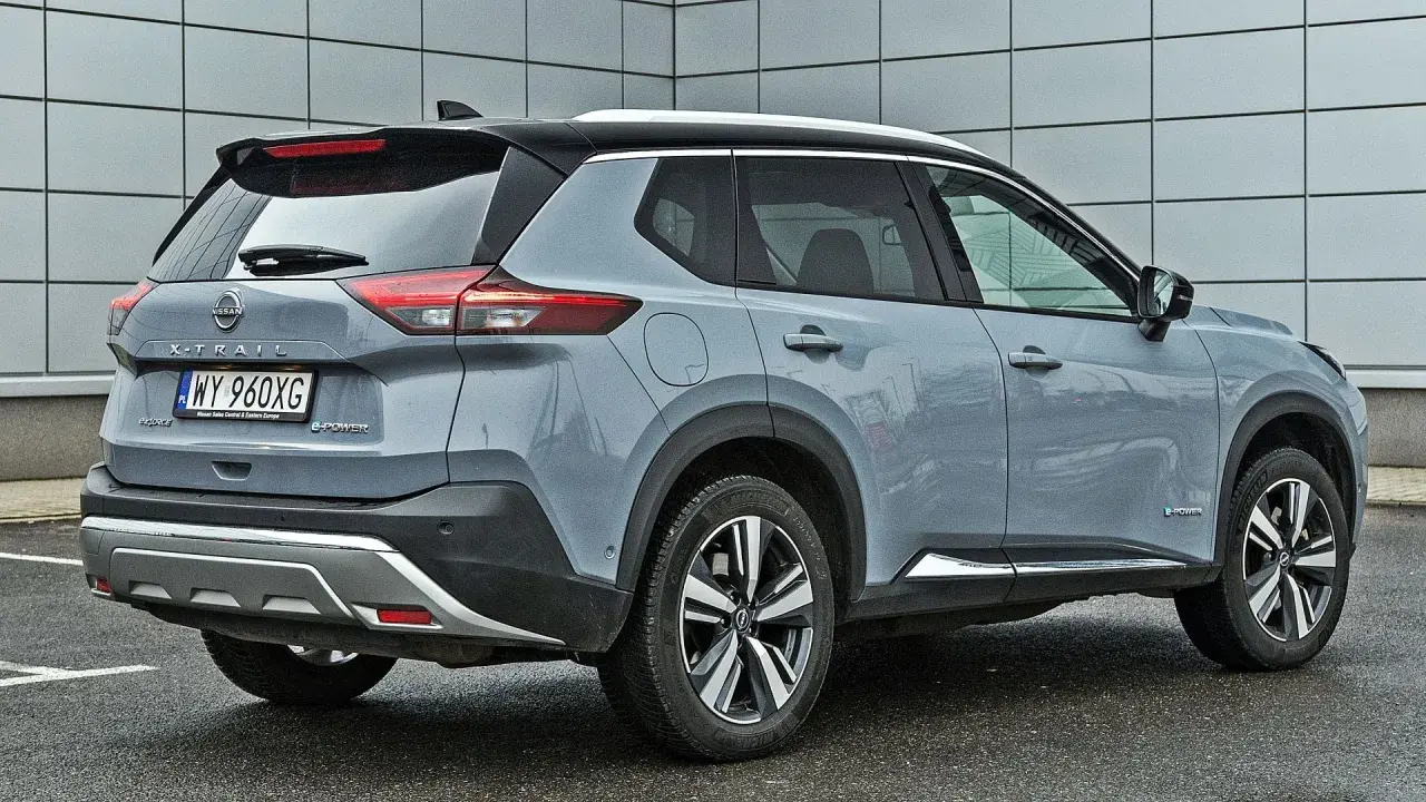 Szary Nissan X-Trail e-POWER z czarnym dachem i srebrnymi akcentami stoi na mokrym parkingu przed nowoczesnym budynkiem.