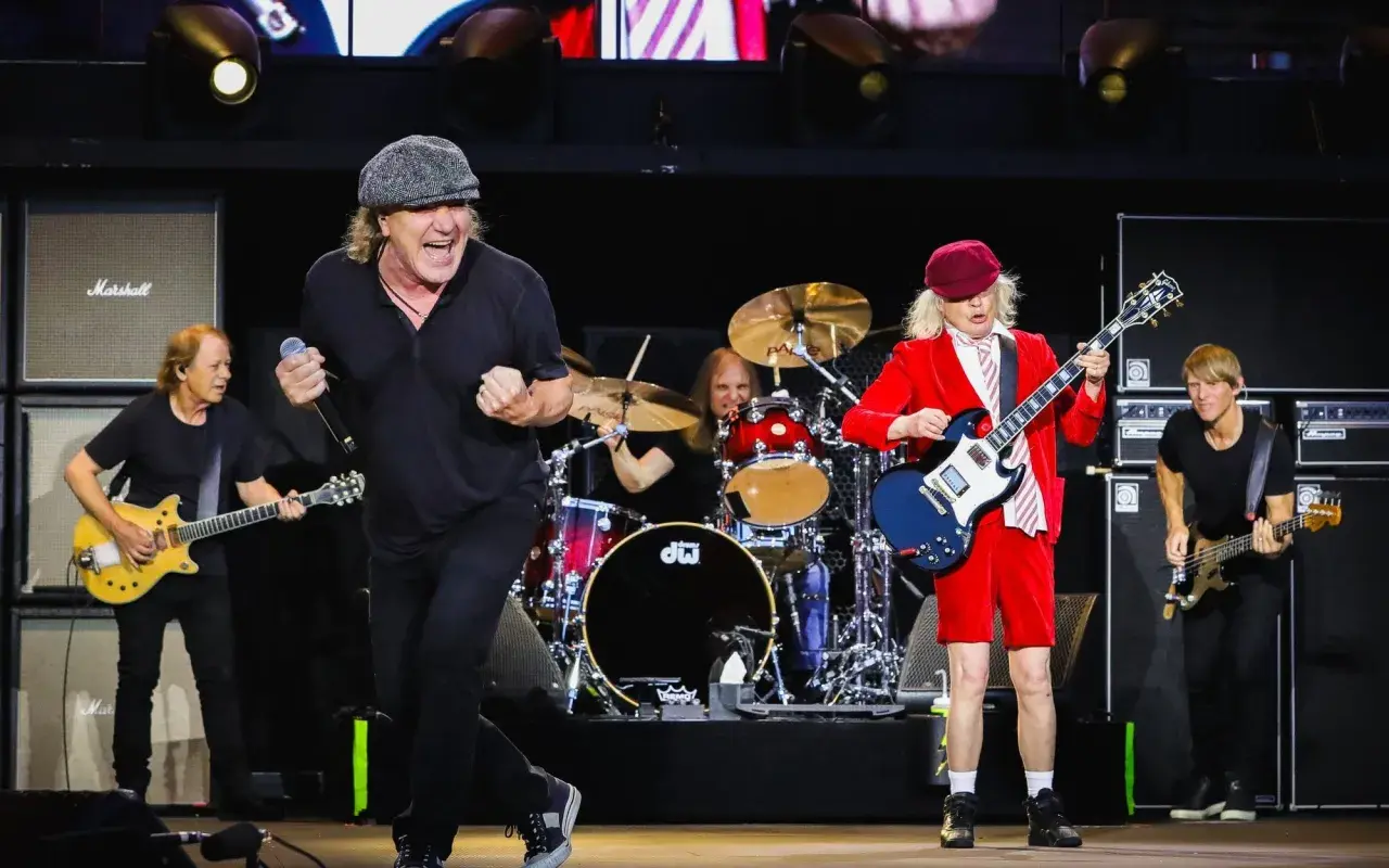 Zespół AC/DC na scenie, grający swoje kultowe utwory. Wokalista w czapce, gitarzysta w czerwonym stroju.