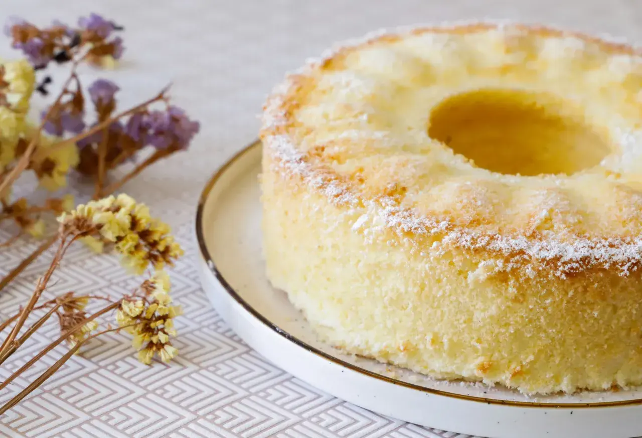 Delizioso chiffon cake ricetta originale americana, leggero e soffice, spolverato di zucchero a velo, con fiori secchi sullo sfondo.