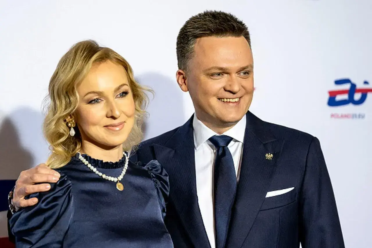 Szymon Hołownia z żoną, uśmiechnięci, pozują do zdjęcia. Ona w granatowej sukience i perłach, on w garniturze.