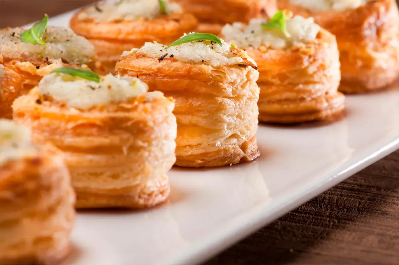 Petites bouchées à la reine dorées, garnies d'une préparation crémeuse et d'une feuille de basilic, présentées sur un plat blanc.