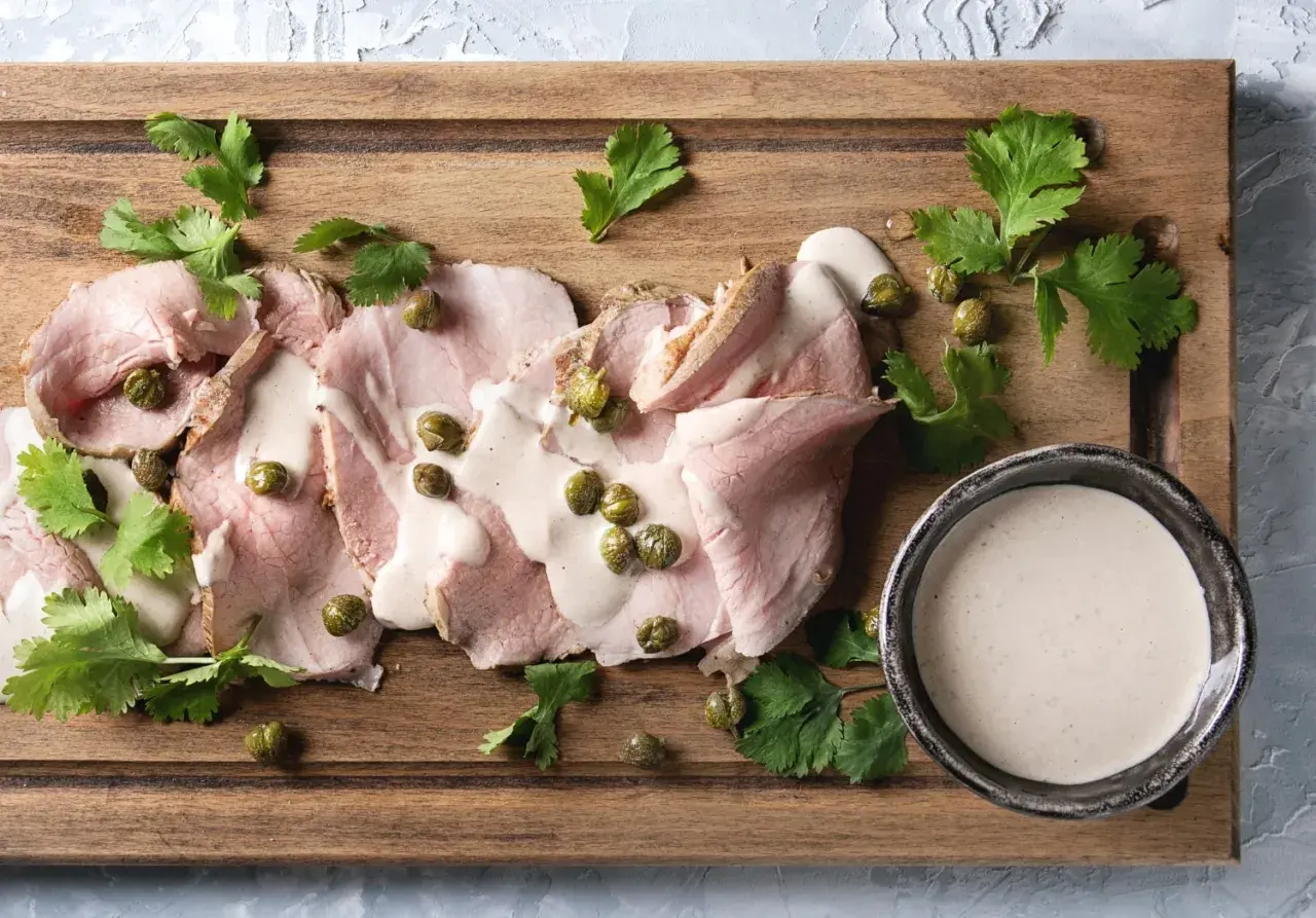 Fette di arrosto con salsa tonnata, capperi e prezzemolo su tagliere di legno.