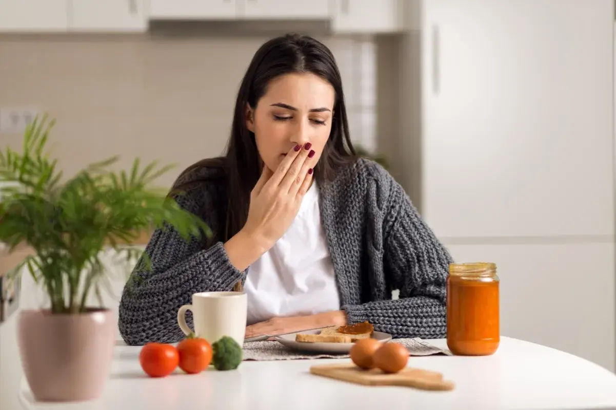 Donna con nausea e debolezza alle gambe si copre la bocca, seduta a tavola con colazione.