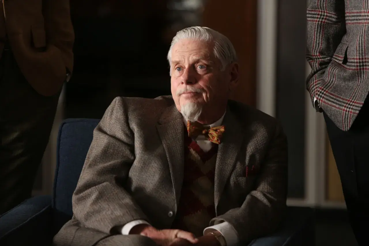 Äldre man med grått hår och bockskägg, klädd i kavaj och fluga. Robert Morse ser fundersam ut.
