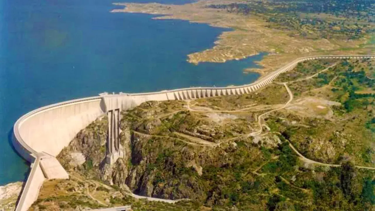 Vista aérea de la imponente presa de Almendra, reteniendo un vasto embalse de agua azul. La estructura curva se alza majestuosa sobre un paisaje rocoso y verde.