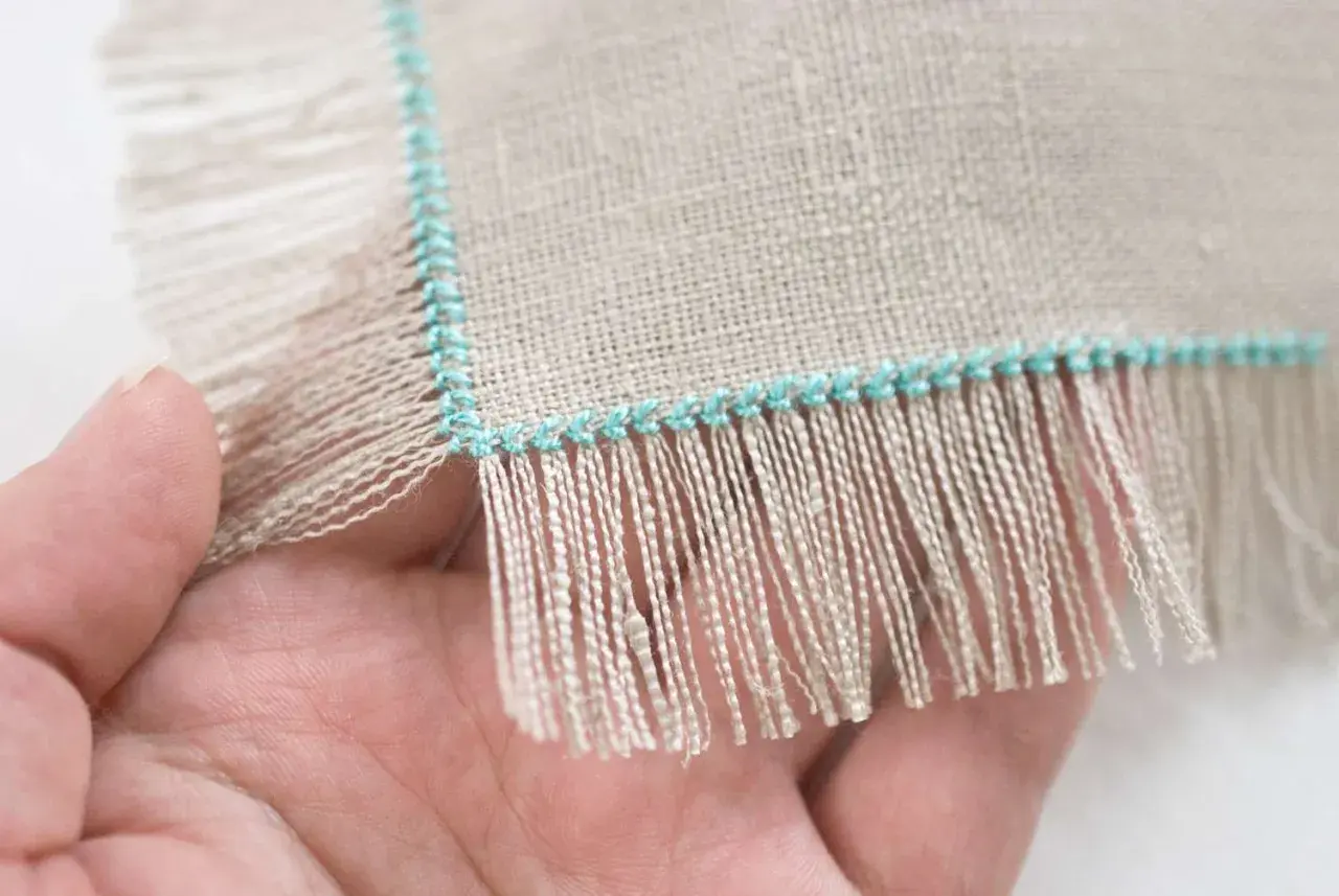 Close-up of a hand holding linen fabric with a turquoise stitched edge, showing how to sew fringe.