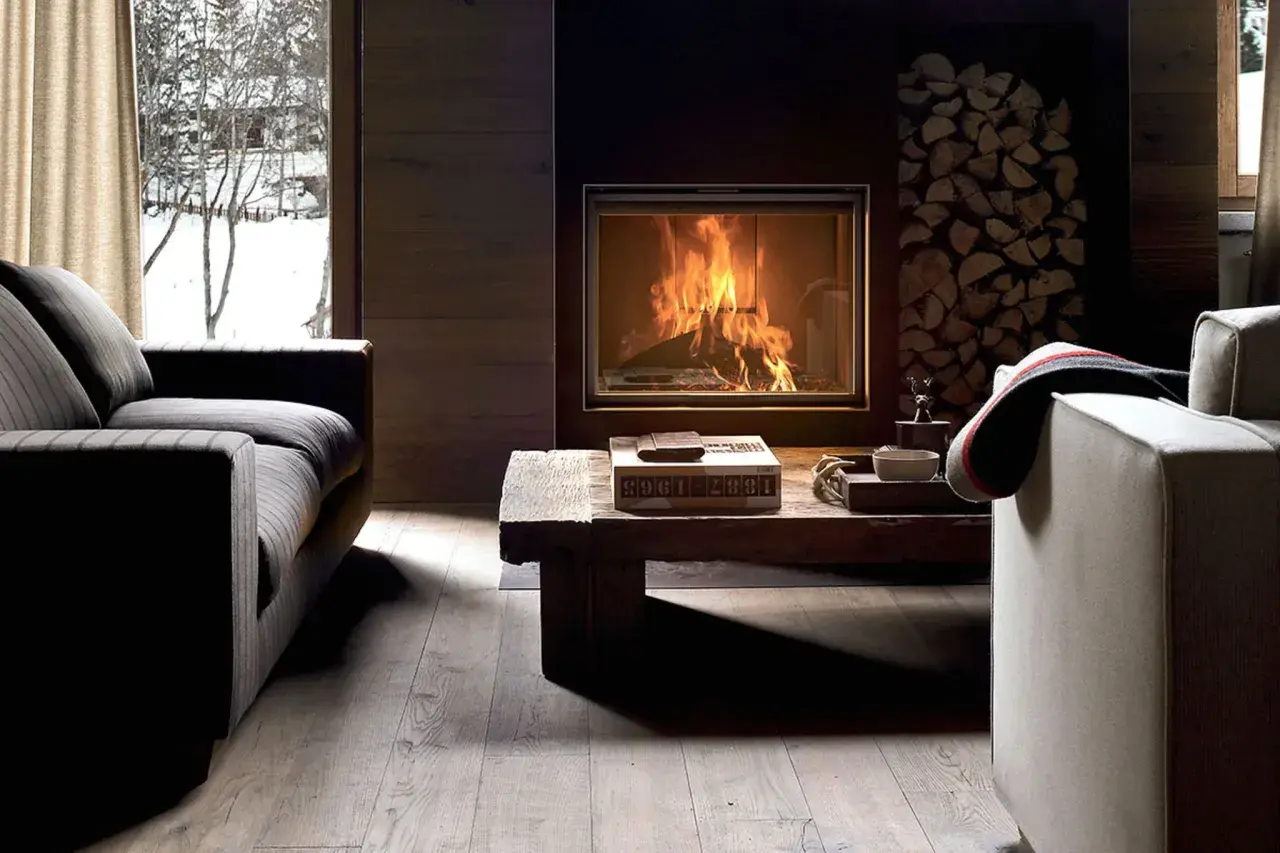 Gemütliches Wohnzimmer mit Kaminfeuer, Holzstapel und Blick auf verschneite Bäume. Tolle Kamin Ideen für Ihr Wohnzimmer.