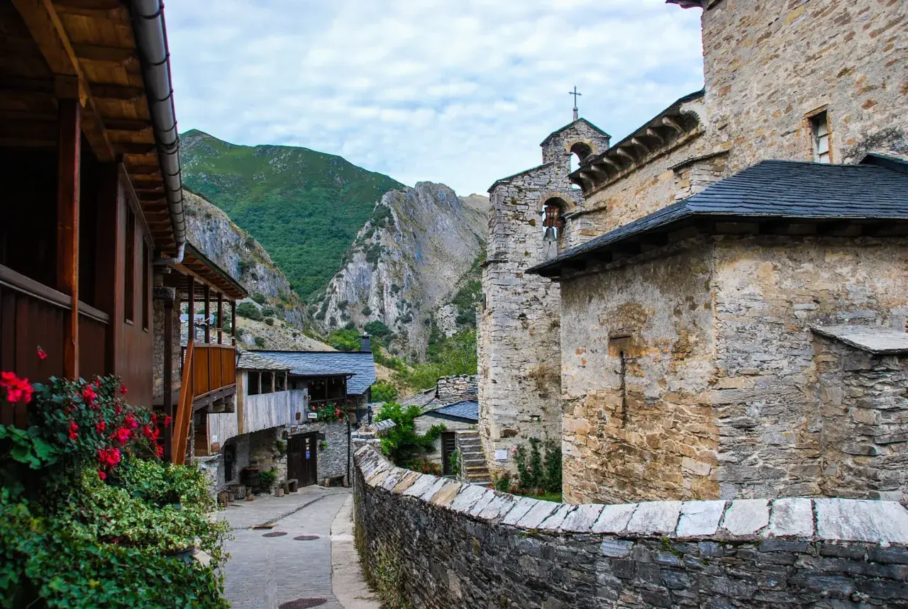 Un pueblo de piedra y madera se alza en el valle del silencio, rodeado de montañas verdes y un cielo nublado.
