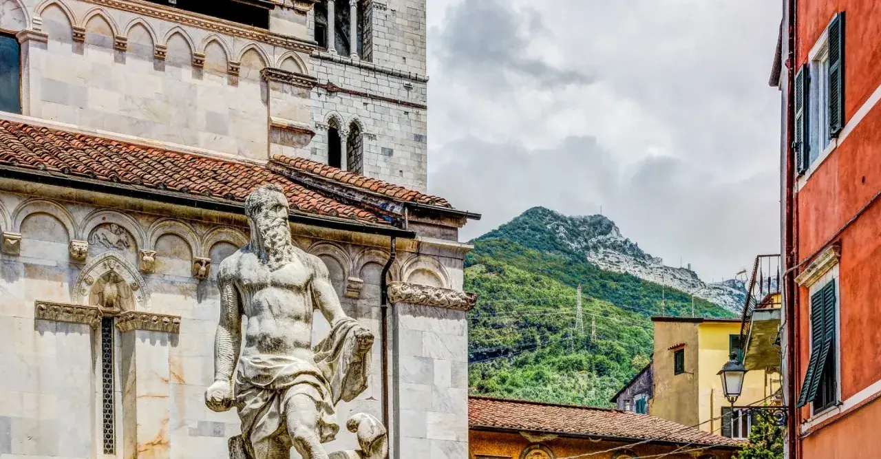Statua nel carrara centro storico, con montagne verdi sullo sfondo e case colorate.