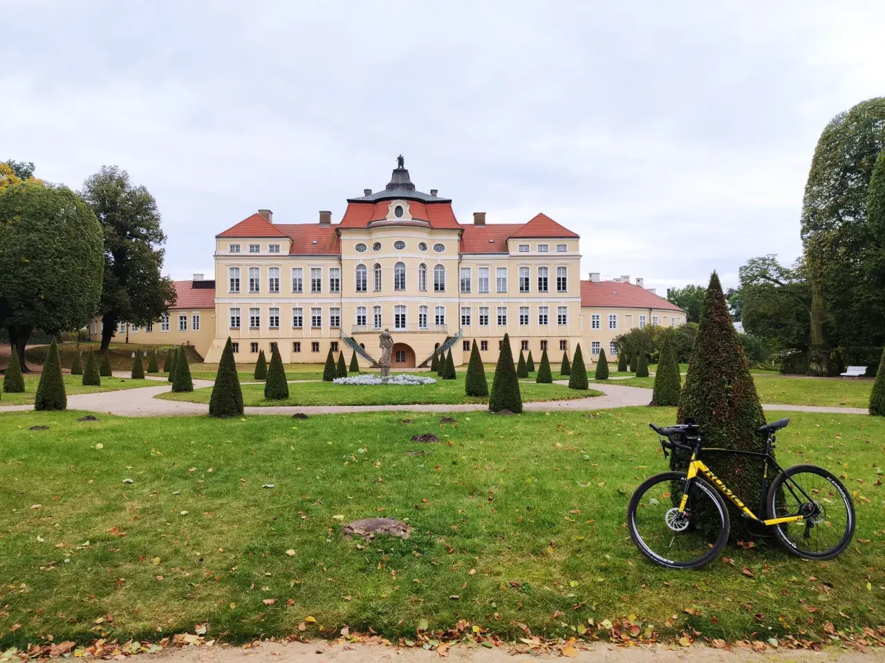 Pałac w Strykowie z żółtą ramą roweru na pierwszym planie, otoczony zadbanym parkiem z rzędami stożkowych drzew.