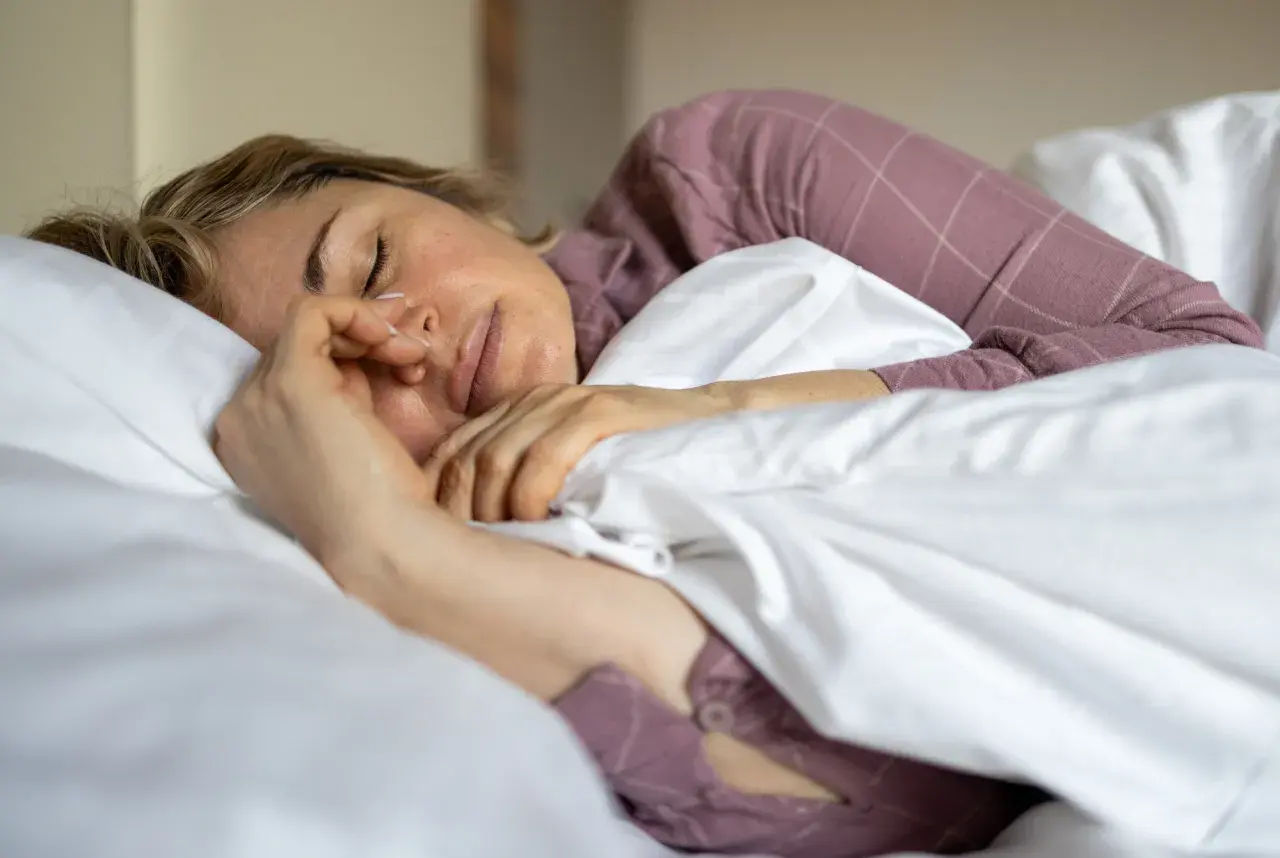 En kvinna sover 6 timmar i en säng med vita lakan och en lila rutig pyjamas. Hon håller handen mot ansiktet.