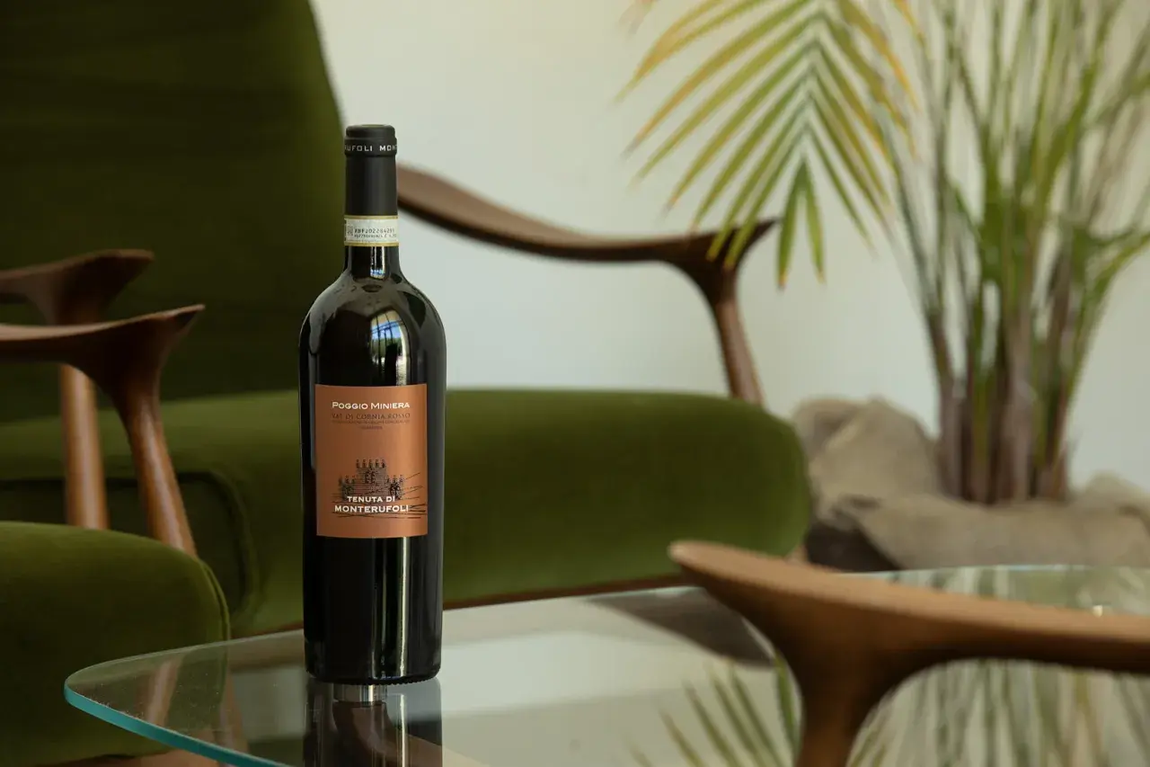 Bottiglia di vino rosso Poggio Miniera, un'eccellenza tra i vini toscani, su un tavolino di vetro, con poltrone verdi e piante sullo sfondo.