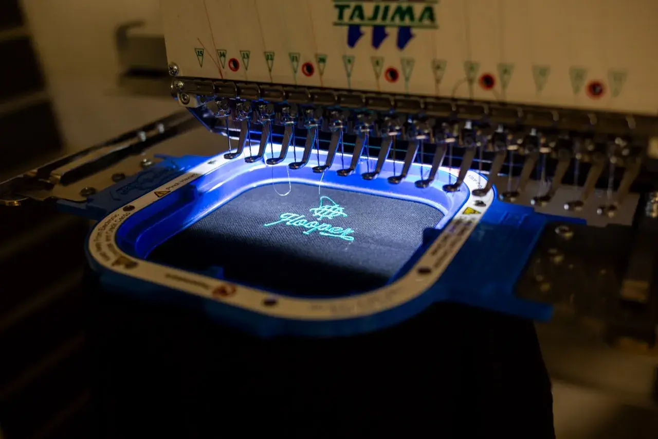 Machine à broder Tajima créant une estampille digitalisée sur un tissu noir. Le logo "Hooper" est en cours de réalisation avec du fil turquoise.