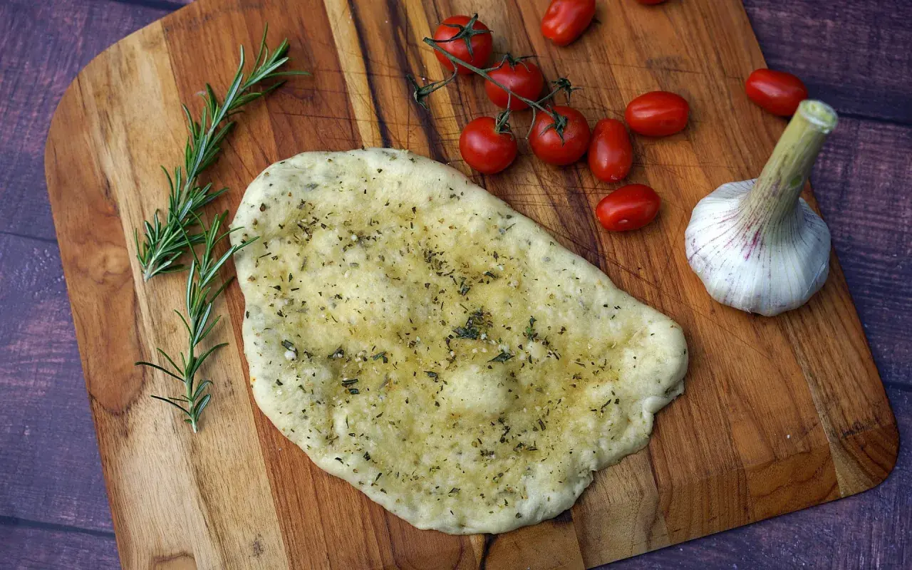 Ein unfertiges Pizzabrot mit Knoblauch und Kräutern liegt auf einem Holzbrett, daneben Kirschtomaten und Rosmarin.