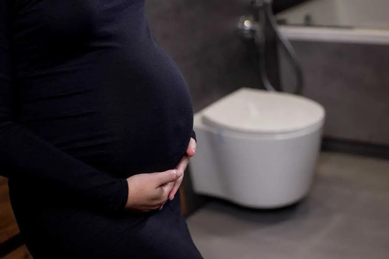 Gravid kvinna med händer på magen, toalett i bakgrunden. Vanliga besvär som uvi kan drabba gravida.