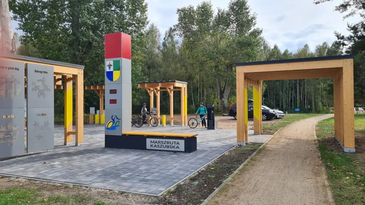 Nowiutka stacja rowerowa na Marszrucie Kaszubskiej. Dwie panie z rowerami, nowoczesna infrastruktura i leśne otoczenie.