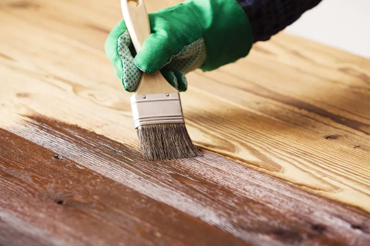 Mano con guante verde aplicando barniz sobre madera, creando un acabado brillante. Barnizar sobre barniz sin lijar para renovar la superficie.