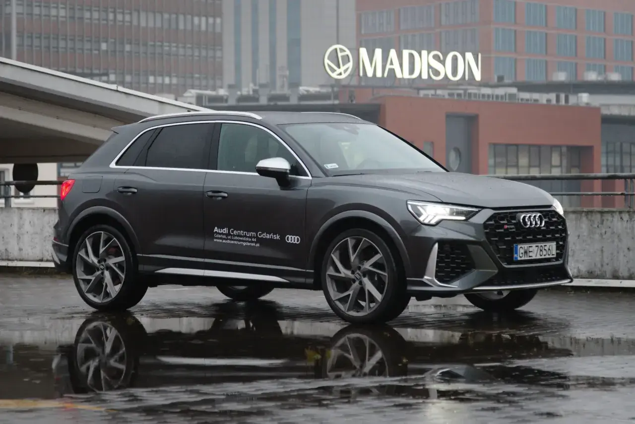 Szary Audi RS Q3 na mokrej nawierzchni, z odbiciem w kałuży. W tle budynek z napisem MADISON.