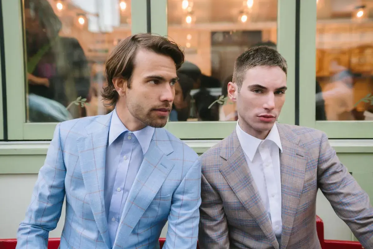 Dwóch mężczyzn w garniturach w kratę. Jeden w błękitnym, drugi w brązowym. Eleganckie klapy w marynarce podkreślają ich styl.
