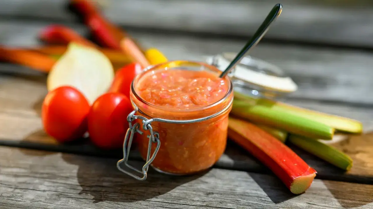 En burk med rabarber chutney står på ett träbord, omgiven av färska rabarber, tomater och en lök.