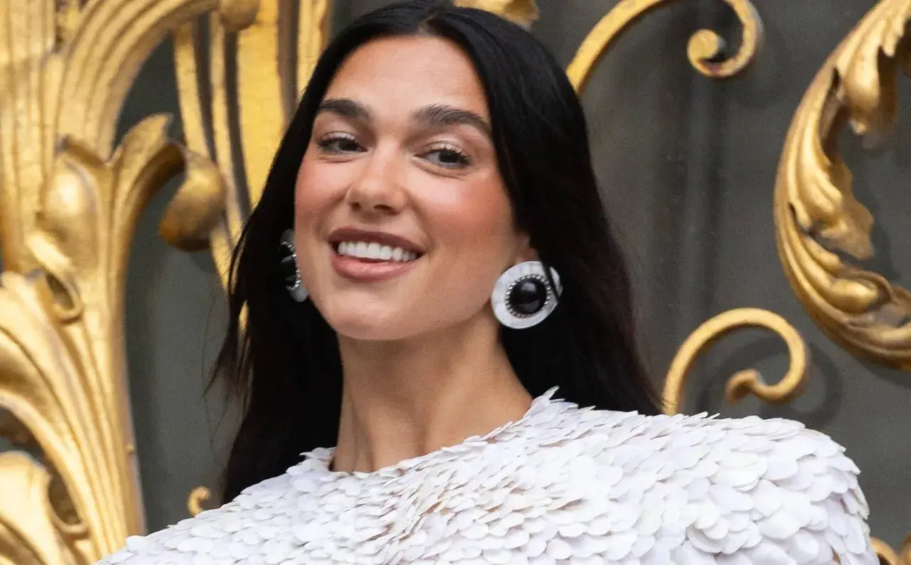 Uśmiechnięta Dua Lipa w białej kreacji, z dużymi kolczykami. Jej albańskie pochodzenie dodaje jej uroku.