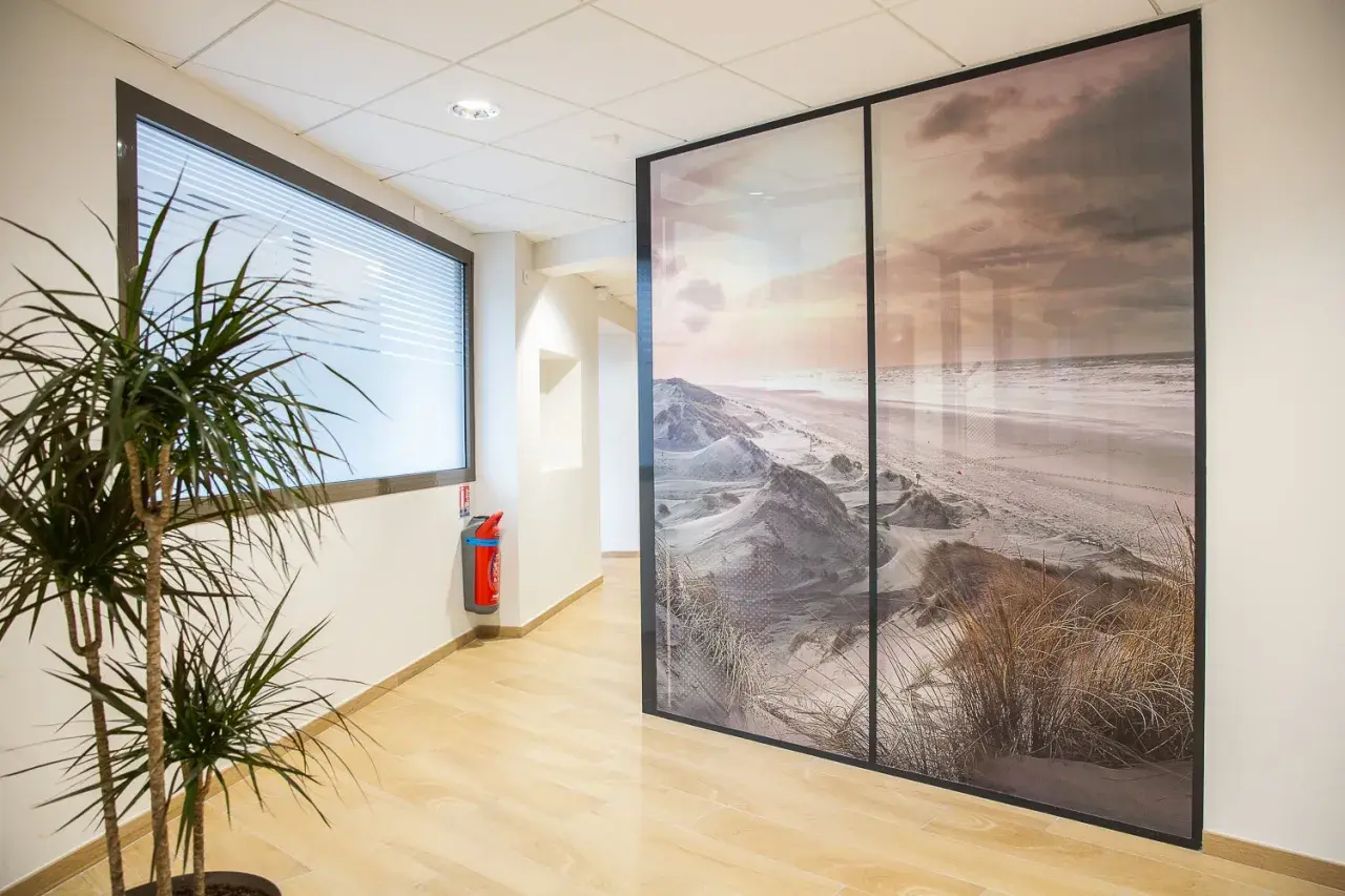 Un couloir moderne avec une plante verte et un extincteur, séparé par une grande cloison amovible ornée d'un paysage de dunes et de plage.