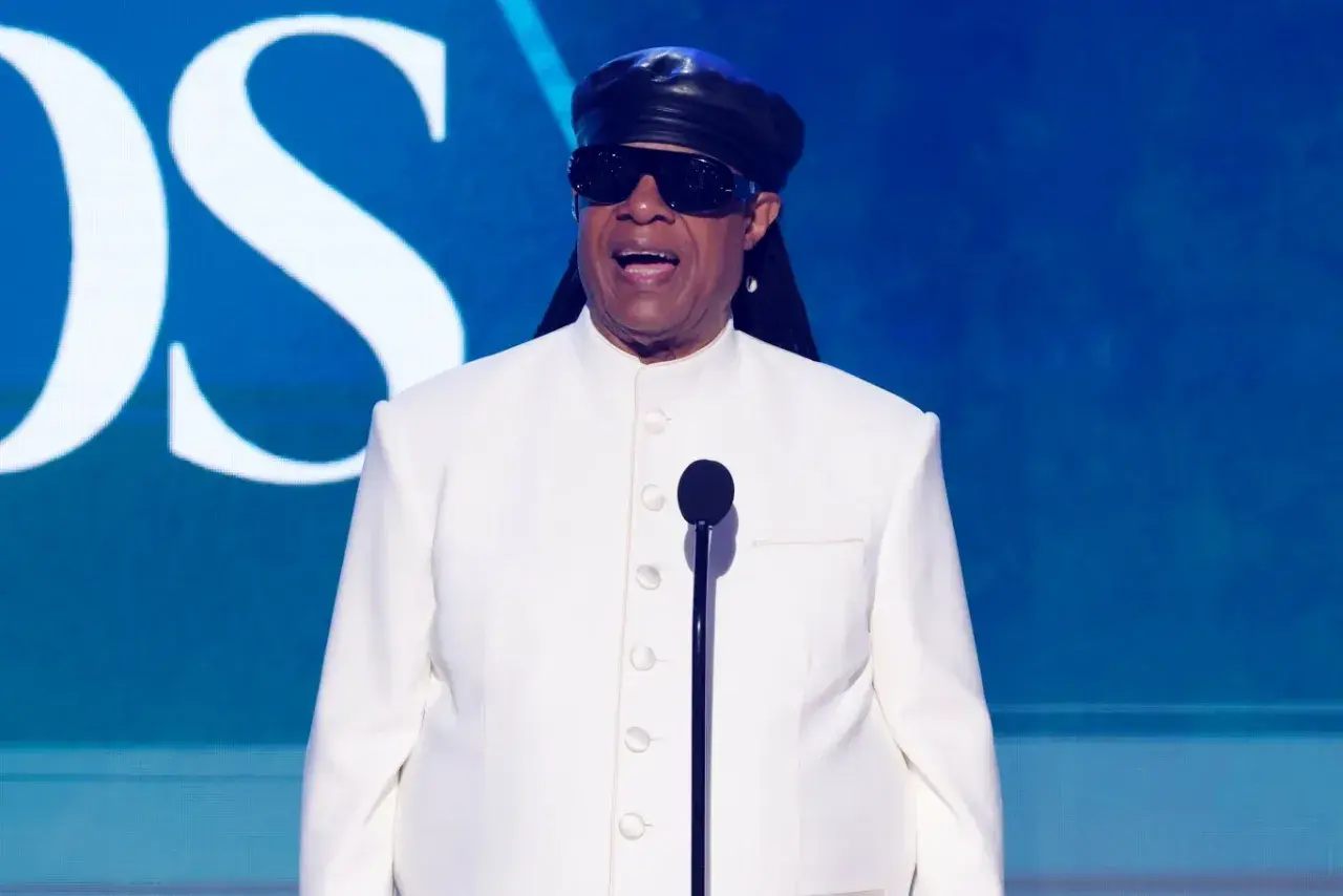 Niewidomy piosenkarz Stevie Wonder w białym garniturze i czarnej czapce, przemawia do mikrofonu na tle niebieskiej sceny z logo CBS.
