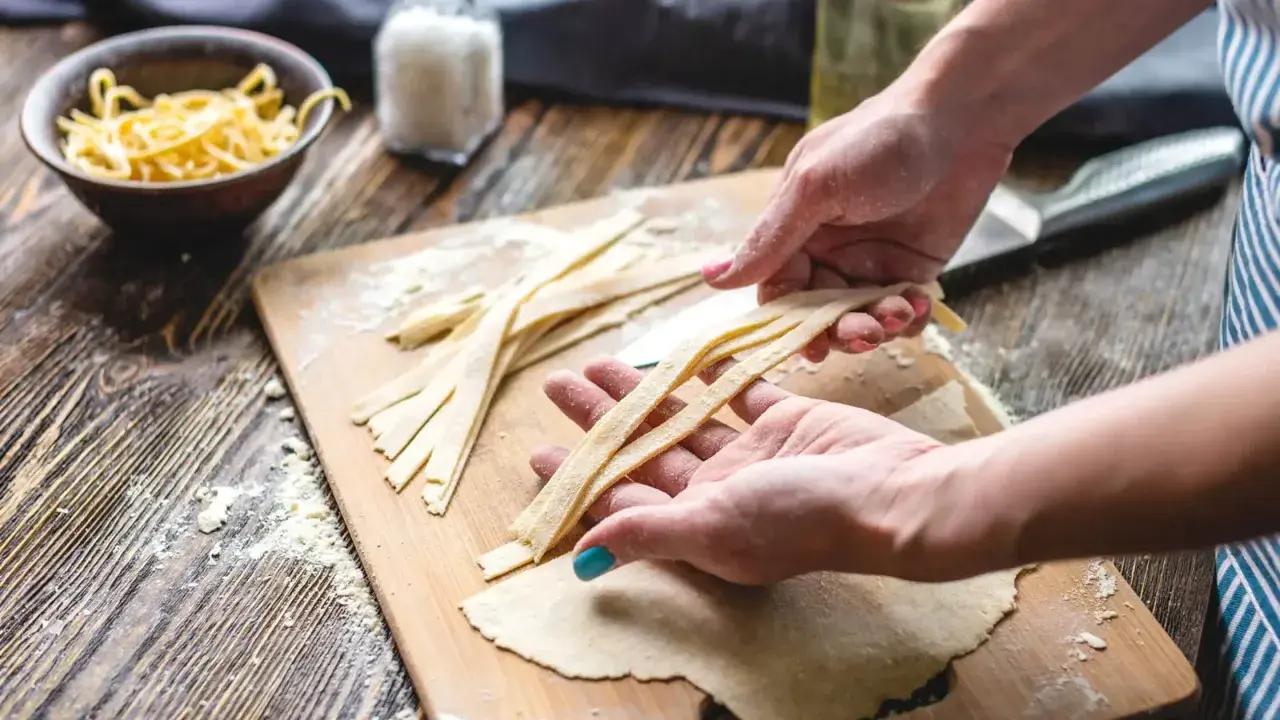 Hände formen frische Pasta-Streifen auf einem Holzbrett mit Mehl.