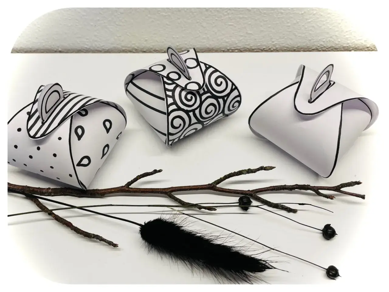 Drei selbstgebastelte Geschenkboxen mit Mustern, inspiriert von Vorlagen zum Geschenkbox selber basteln. Ein Ast und schwarze Federn liegen daneben.