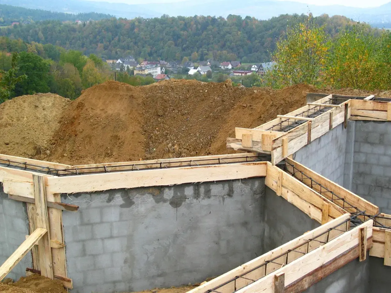 Fundament budowy z szalunkiem i zbrojeniem, w tle domy i las.