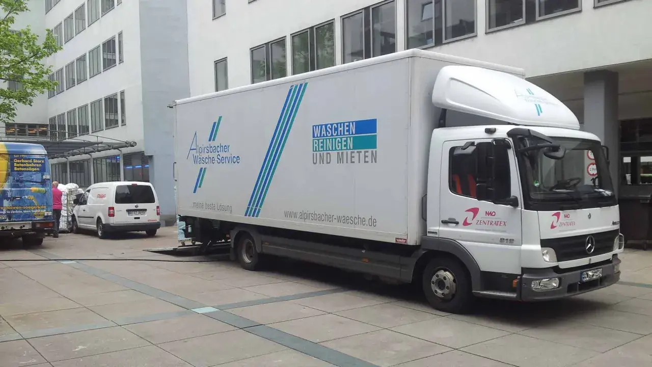 Ein LKW von Alpirsbacher Wäsche Service, der für Werkverkehr genutzt wird, steht vor einem Gebäude. Er transportiert Wäsche.