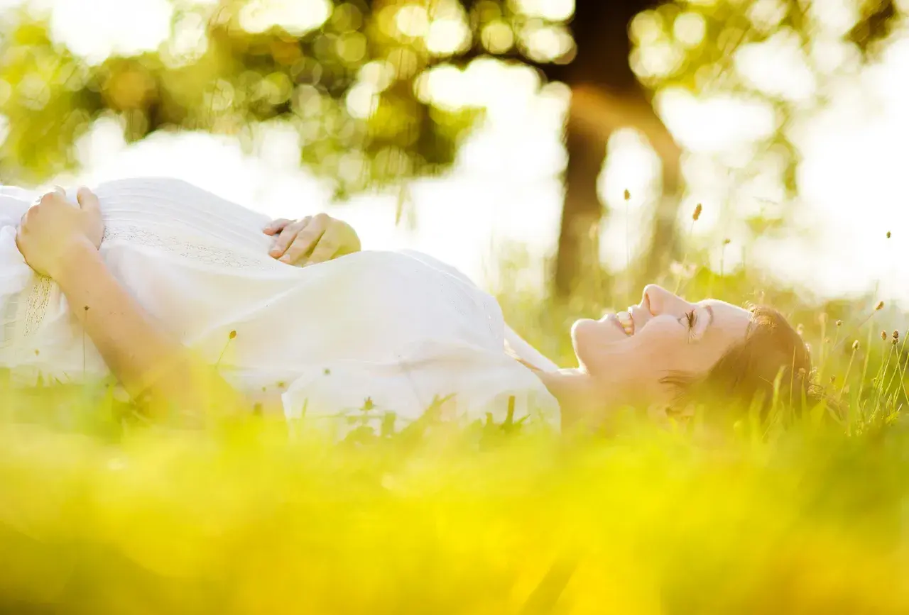 Une femme enceinte, baignée par le soleil, sourit en se prélassant dans l'herbe verte.