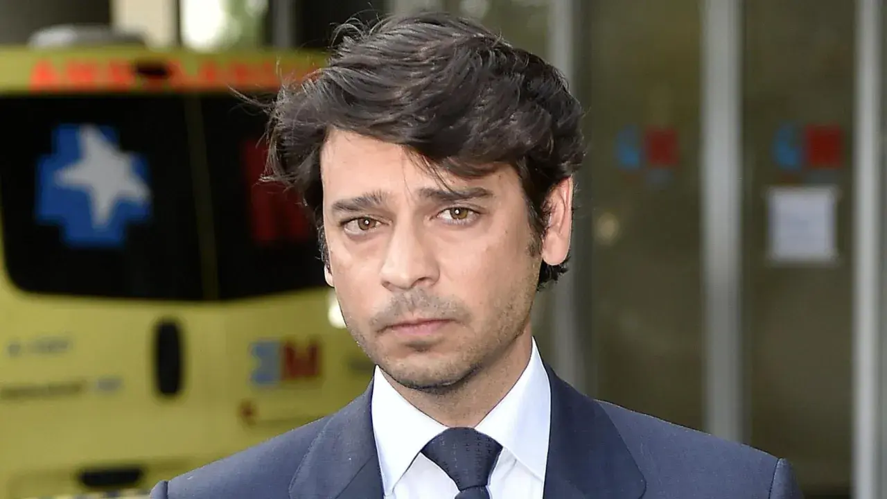 Miguel Palomo Danko, con traje oscuro y corbata, mira fijamente. Detrás, una ambulancia amarilla.