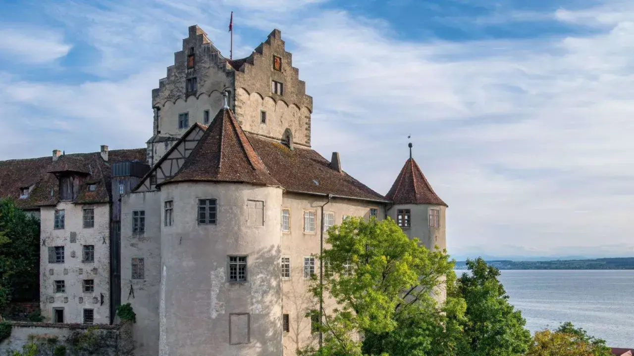 Das Meersburg Castle thront über dem Bodensee, umgeben von grünen Bäumen und unter einem blauen Himmel mit Wolken.