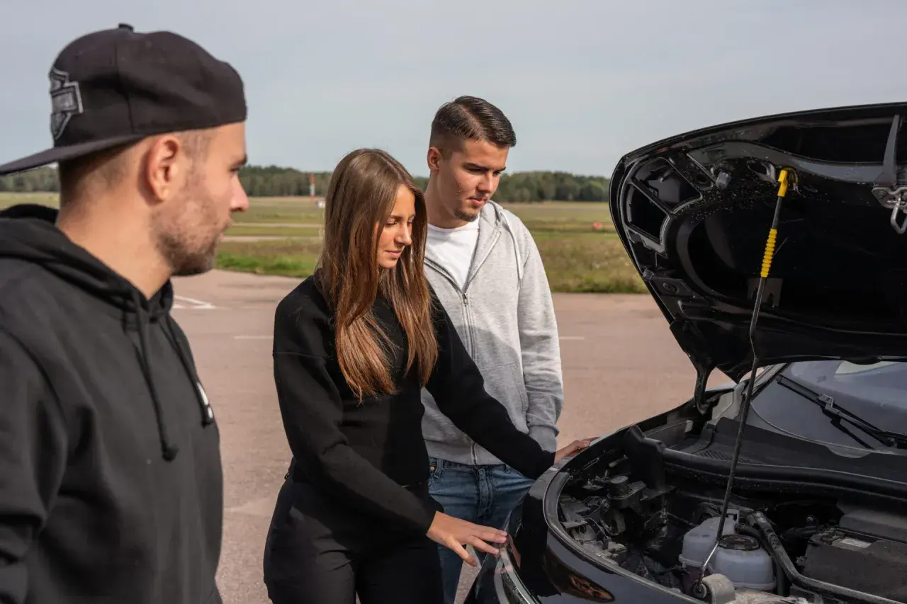 Tre unga personer tittar på en bil med öppen motorhuv. Kanske behöver de hjälp av en handledare bil.